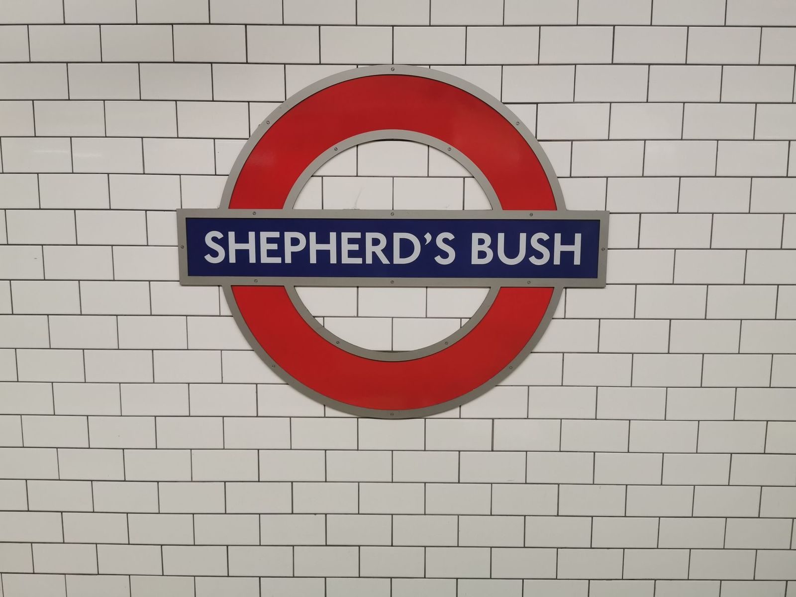 A red Underground roundel reading Shepherd's Bush on a white tiled wall