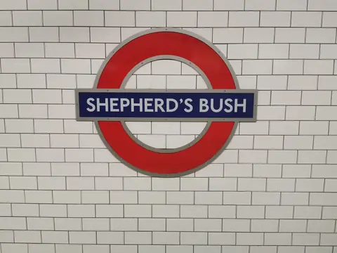 A red Underground roundel reading Shepherd's Bush on a white tiled wall